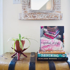 Może przedstawiać: Dekoracyjna ekspozycja z książkami o tytułach "Global Style", "Bohemian Style" i "Heirloom Modern". Roślina doniczkowa stoi na dekoracyjnym stojaku. Rzeźbione lustro wisi nad ekspozycją.