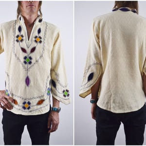 May include: Cream-coloured long-sleeve shirt with colourful embroidered floral and geometric patterns. The shirt has a V-neck and flared sleeves, with decorative stitching around the collar and cuffs. The shirt is made of a textured fabric.