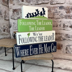 May include: Four rectangular wooden signs stacked on a small table. The top sign is white with a black silhouette of characters. The other signs are green, white, and navy blue, each with white text that reads "Following The Leader."