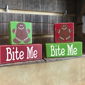 May include: Wooden blocks with a gingerbread man design and the text "Bite Me". The blocks are painted in red and green with white lettering. The gingerbread men are brown with white icing details.
