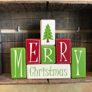 May include: Wooden blocks spelling "MERRY Christmas" and a Christmas tree, arranged inside a rustic wooden crate. The blocks are painted in red, green, and white, creating a festive holiday decoration.