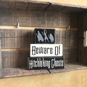 May include: Three rectangular wooden signs with a spooky theme. The top sign is black with gray silhouettes. The middle sign is white with the words "Beware Of" in black. The bottom sign is black with "Hitchhiking Ghosts" in white.