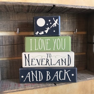 May include: A stack of four wooden blocks with white lettering. The top block is navy blue with a white moon, stars, and silhouettes of Peter Pan and the Darling children. The next block is green with "I LOVE YOU" in white. The third block says "TO NEVERLAND" and the bottom block says "AND BACK".