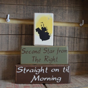 May include: Three wooden blocks with painted text and images. The top block features a black silhouette of a ship against a yellow background. The middle block reads "Second Star from The Right." The bottom block says "Straight on til Morning."
