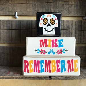May include: A Day of the Dead themed display featuring a black square block with a skull design and two rectangular blocks. The rectangular blocks read "MIKE" and "REMEMBER ME" in colorful letters. The background is a wooden shelf.
