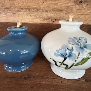 May include: Two ceramic oil lamps. One is a solid blue, the other is white with blue floral accents. Both have a round body, a small neck, and a wick. They are sitting on a wooden surface.