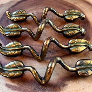May include: Four antique brass leaf-shaped drawer pulls arranged on a wooden surface. Each pull features a stylized leaf design with a curved stem and a central, zig-zag handle. The brass has a warm, aged patina.