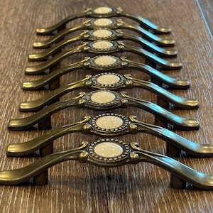 May include: A set of eight antique-style cabinet pulls. Each pull has a curved, bronze-toned handle with a decorative, oval center featuring a cream-colored, speckled insert. The pulls are arranged in parallel rows on a wooden surface.