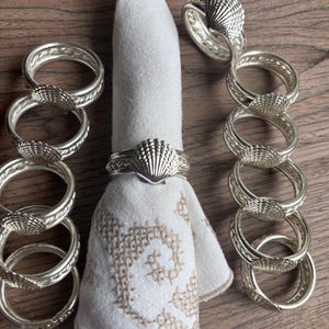 May include: A collection of silver-toned seashell napkin rings. The rings are arranged around a white napkin with a beige embroidered design. The rings have a detailed seashell design in the center.