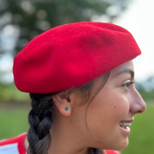 May include: A bright red beret made of a soft, felt-like material. The beret is a classic style, round with a flat crown. The hat is worn at an angle, and is a solid, vibrant color. The beret is a stylish accessory.