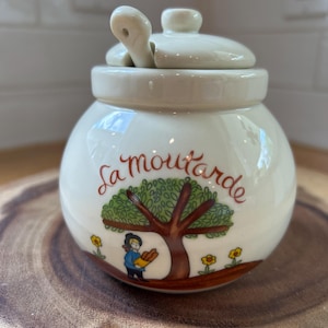 May include: Cream-colored ceramic mustard pot with a lid and a small spoon. The pot features a tree illustration with the words "La moutarde" in red script. A person is depicted holding bread.