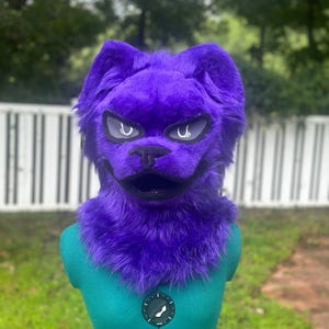 May include: A purple faux fur animal head mask featuring pointed ears, a black nose, and an open mouth. The mask has stylized, sharp-angled eyes with white pupils and dark outlines, set against a vibrant, deep purple textured fur covering.