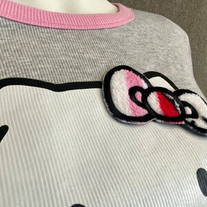 May include: Gray long-sleeved shirt with a pink ribbed neckline and a white Hello Kitty appliqu&eacute; with a pink and red bow.