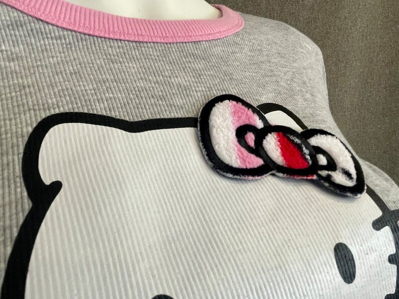 May include: Gray long-sleeved shirt with a pink ribbed neckline and a white Hello Kitty appliqu&eacute; with a pink and red bow.