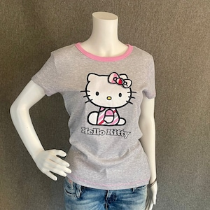 May include: Gray short-sleeved t-shirt with a pink trim around the neck. The shirt features a Hello Kitty graphic with a pink and white striped dress and a pink bow.