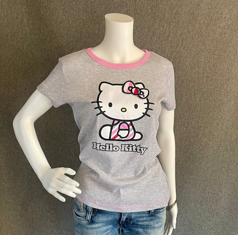 May include: Gray short-sleeved t-shirt with a pink trim around the neck. The shirt features a Hello Kitty graphic with a pink and white striped dress and a pink bow.