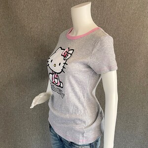May include: Gray short-sleeved t-shirt with a pink trim around the neckline. The shirt features a Hello Kitty graphic with a pink bow.