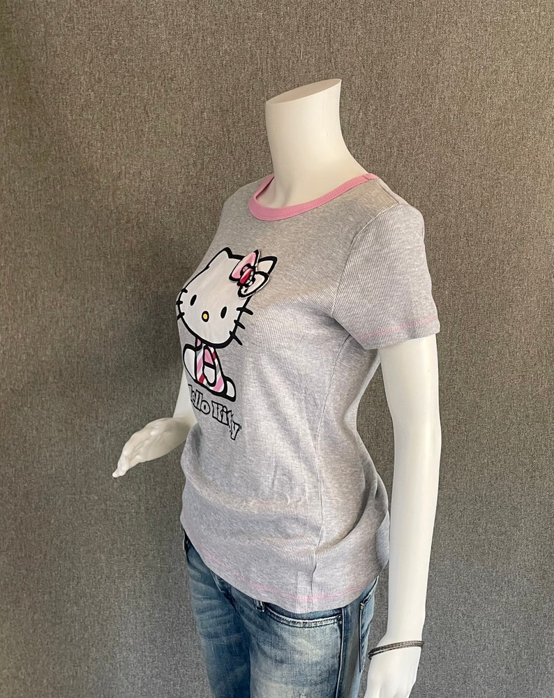 May include: Gray short-sleeved t-shirt with a pink trim around the neckline. The shirt features a Hello Kitty graphic with a pink bow.
