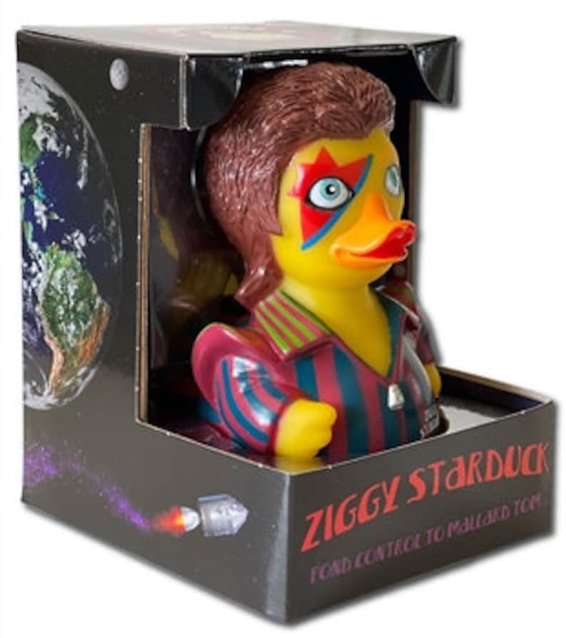 Peut inclure: Un canard en plastique dans une bo&icirc;te, stylis&eacute; comme Ziggy Stardust, avec une chemise ray&eacute;e rouge et bleue et du maquillage. La bo&icirc;te pr&eacute;sente un th&egrave;me spatial avec une image de la Terre et le texte "ZIGGY STARDUCK".