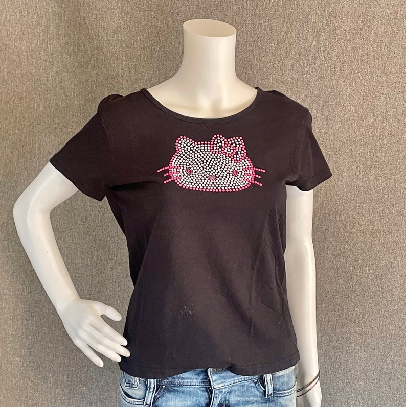 Puede incluir: Camiseta negra con un dise&ntilde;o de Hello Kitty de pedrer&iacute;a. El dise&ntilde;o presenta una cara de Hello Kitty blanca con detalles rosas.