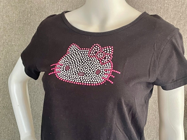 Puede incluir: Camiseta negra con un dise&ntilde;o de Hello Kitty rosa y blanco hecho de pedrer&iacute;a.