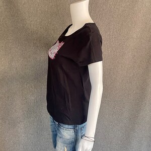 Puede incluir: Camiseta negra de manga corta con un dise&ntilde;o gr&aacute;fico rosa y blanco en la parte delantera. El dise&ntilde;o gr&aacute;fico es una imagen estilizada de una ola.