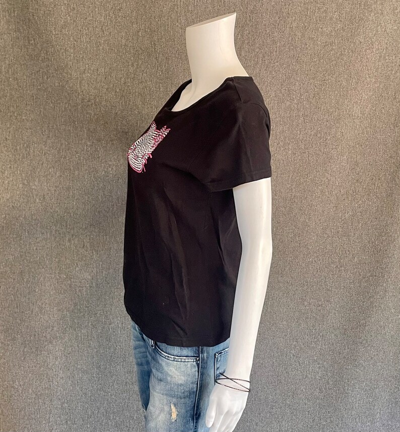 Puede incluir: Camiseta negra de manga corta con un dise&ntilde;o gr&aacute;fico rosa y blanco en la parte delantera. El dise&ntilde;o gr&aacute;fico es una imagen estilizada de una ola.