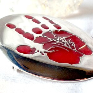 May include: A silver oval-shaped pendant with a red handprint design. The handprint is raised and has a textured surface.