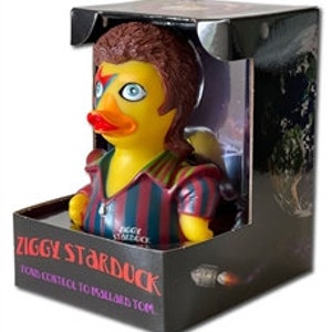 Peut inclure: Un canard en plastique sur le th&egrave;me de Ziggy Stardust, pr&eacute;sent&eacute; dans une bo&icirc;te noire. Le canard a un corps jaune, des yeux bleus et des l&egrave;vres rouges. Il porte une chemise ray&eacute;e et a les cheveux bruns. La bo&icirc;te indique "ZIGGY STARDUCK" et "TOMS CONTROL TO MALLARD TOM..."