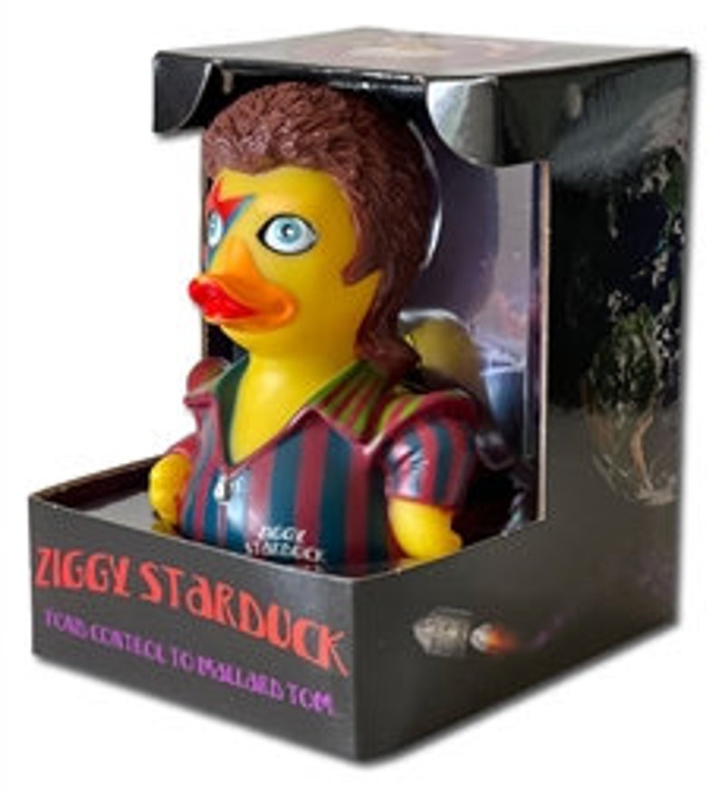 Peut inclure: Un canard en plastique sur le th&egrave;me de Ziggy Stardust, pr&eacute;sent&eacute; dans une bo&icirc;te noire. Le canard a un corps jaune, des yeux bleus et des l&egrave;vres rouges. Il porte une chemise ray&eacute;e et a les cheveux bruns. La bo&icirc;te indique "ZIGGY STARDUCK" et "TOMS CONTROL TO MALLARD TOM..."