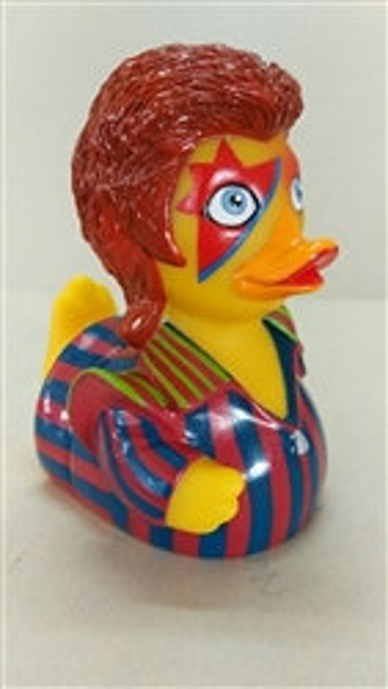 Peut inclure: Un canard en plastique jaune avec un bec orange et des yeux bleus. Le canard est stylis&eacute; pour ressembler &agrave; un musicien c&eacute;l&egrave;bre, avec des cheveux roux, une marque en forme d'&eacute;toile sur le visage et une tenue ray&eacute;e bleue, rouge et or.