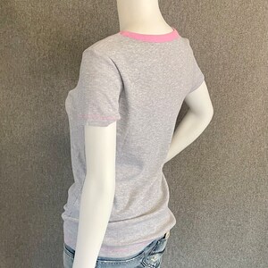 May include: A gray ribbed t-shirt with a pink trim around the neckline and sleeves. The shirt is on a mannequin and is being worn with blue jeans.