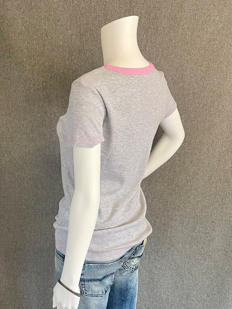 May include: A gray ribbed t-shirt with a pink trim around the neckline and sleeves. The shirt is on a mannequin and is being worn with blue jeans.