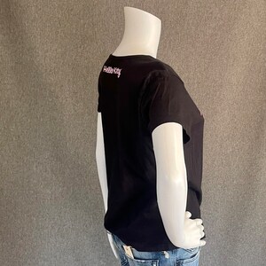 Puede incluir: Camiseta negra de manga corta con un gr&aacute;fico rosa de Hello Kitty en la espalda. La camiseta est&aacute; siendo usada por un maniqu&iacute;.