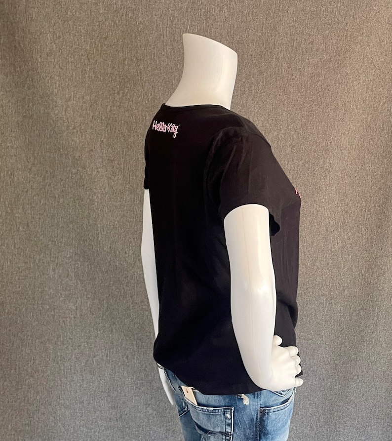Puede incluir: Camiseta negra de manga corta con un gr&aacute;fico rosa de Hello Kitty en la espalda. La camiseta est&aacute; siendo usada por un maniqu&iacute;.