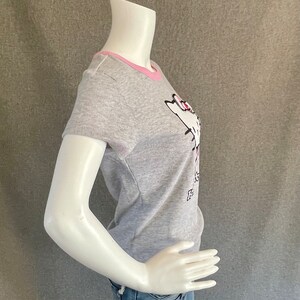 May include: Gray short-sleeved t-shirt with a pink trim around the neckline. The shirt features a white cartoon cat with pink accents and the text "It's a Cat Thing" in black.