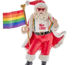 May include: A decorative Santa Claus ornament in a red suit with white fur trim, wearing sunglasses and holding a rainbow flag. The ornament has the words "Love is Love" on the shirt. The belt buckle is heart-shaped.