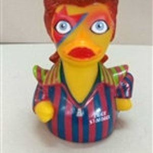 Peut inclure: Un canard en plastique jaune avec des cheveux roux et un &eacute;clair peint sur le visage. Le canard porte une chemise ray&eacute;e avec des rayures rouges et bleues. Le canard a les yeux bleus et un bec rouge.
