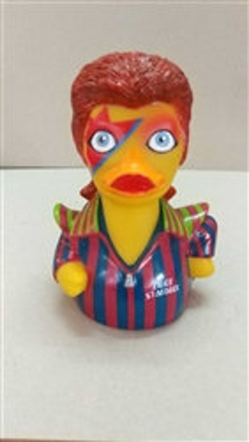 Peut inclure: Un canard en plastique jaune avec des cheveux roux et un &eacute;clair peint sur le visage. Le canard porte une chemise ray&eacute;e avec des rayures rouges et bleues. Le canard a les yeux bleus et un bec rouge.