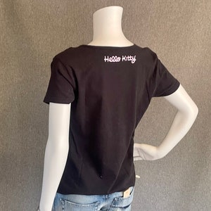 Puede incluir: Camiseta negra de manga corta con un gr&aacute;fico blanco de "Hello Kitty" en la espalda. La camiseta se est&aacute; modelando en un maniqu&iacute; blanco que lleva jeans azules.