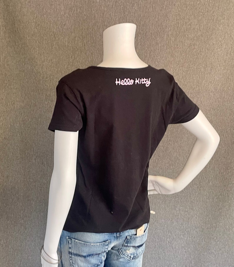 Puede incluir: Camiseta negra de manga corta con un gr&aacute;fico blanco de "Hello Kitty" en la espalda. La camiseta se est&aacute; modelando en un maniqu&iacute; blanco que lleva jeans azules.