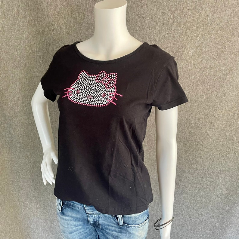 Puede incluir: Camiseta negra con un dise&ntilde;o de Hello Kitty de pedrer&iacute;a. El dise&ntilde;o presenta acentos rosados en el lazo y los bigotes.