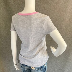 May include: Gray short-sleeved t-shirt with a pink ribbed trim around the neckline.