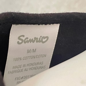 Puede incluir: Etiqueta blanca con texto negro que dice "Sanrio", "M/M", "100% COTTON/COTON", "MADE IN HONDURAS", "FABRIQUE AU HONDURAS", "FSC#30116" y "RN".