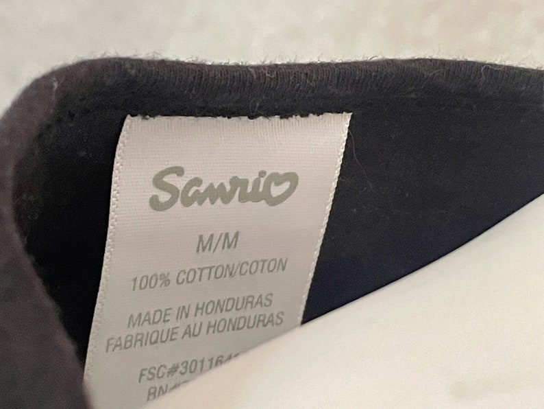 Puede incluir: Etiqueta blanca con texto negro que dice "Sanrio", "M/M", "100% COTTON/COTON", "MADE IN HONDURAS", "FABRIQUE AU HONDURAS", "FSC#30116" y "RN".