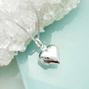 May include: A silver heart pendant necklace. The pendant, a polished heart, hangs from a fine chain. The necklace is presented on a light blue surface, with a white, crystalline backdrop.