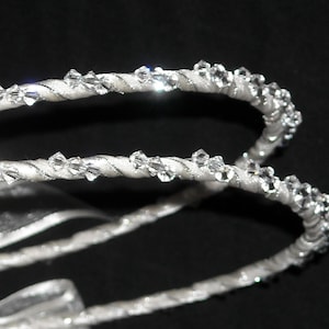 May include: Two white satin-wrapped headbands featuring a twisted cord design adorned with small, clear, faceted crystals that catch the light. The headbands are curved and positioned against a dark background, showcasing the reflective crystal details.