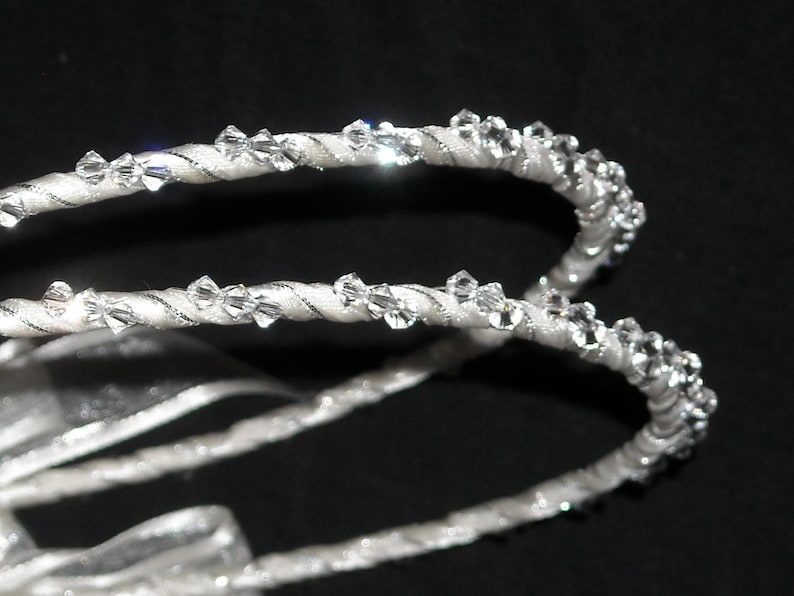 May include: Two white satin-wrapped headbands featuring a twisted cord design adorned with small, clear, faceted crystals that catch the light. The headbands are curved and positioned against a dark background, showcasing the reflective crystal details.
