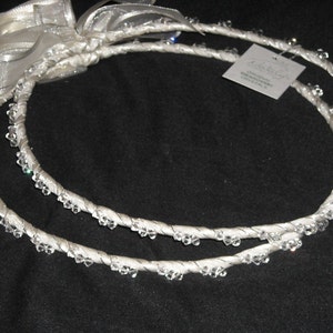 May include: Two circular white crowns wrapped in satin ribbon and adorned with clear Swarovski crystals. The crowns are connected by a white sheer ribbon bow. A small square tag attached to one crown reads Original Swarovski Crystals.