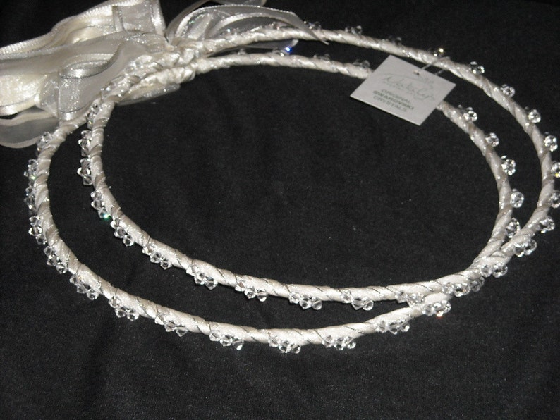 May include: Two circular white crowns wrapped in satin ribbon and adorned with clear Swarovski crystals. The crowns are connected by a white sheer ribbon bow. A small square tag attached to one crown reads Original Swarovski Crystals.